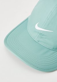 Světle modrá kšiltovka s zakřiveným špichem, vyrobená z hladké tkaniny. Na přední straně se nachází bílé logo Nike a viditelné detaily šití.