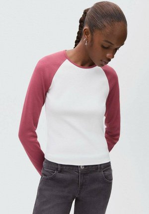 Jeune femme aux cheveux tressés portant une chemise blanche avec des manches raglan longues roses et un jean gris foncé, regardant vers le bas.
