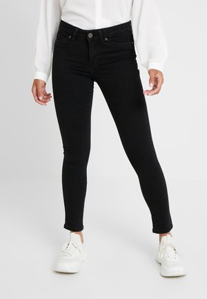 Person wearing fitted black jeans, white long-sleeve top, and white sneakers standing against a plain background.