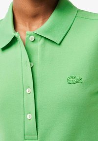 Polo vert en tissu texturé, doté d'une patte de boutonnage à trois boutons et d'un petit logo vert brodé sur le côté gauche de la poitrine.