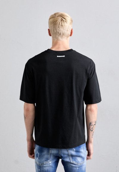 Persona bionda di spalle che indossa una T-shirt nera Dsquared2 e jeans blu strappati, con tatuaggi visibili su entrambi i avambracci.