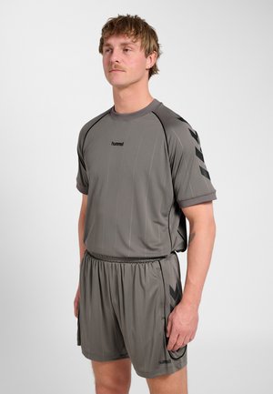 Mężczyzna stojący w dopasowanym szarym komplecie sportowym z logo hummel i wzorem jodełki na rękawach i szortach.