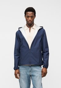 Άνδρας που φοράει ανοιχτό μπουφάν με κουκούλα σε ναυτικό μπλε πάνω από μπεζ πόλο με φερμουάρ και ανοιχτό γαλάζιο τζιν, στέκεται μπροστά σε απλό φόντο.