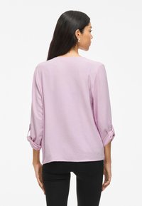Blusa lilla con tessuto testurizzato, maniche a tre quarti arrotolate, scollatura tonda e vestibilità rilassata, abbinata a pantaloni neri.