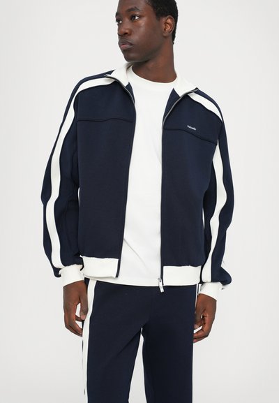 Navy zippered jacket with white side stripes, ribbed cuffs, and collar, paired with matching navy pants. Material appears soft and lightweight.