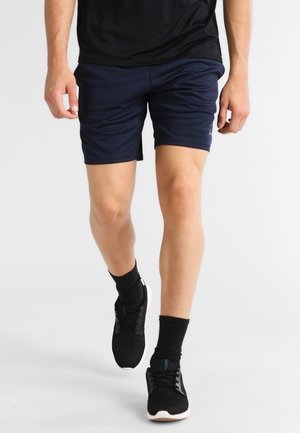 Ein Mann läuft, trägt marineblaue Sportshorts, schwarze Kniestrümpfe, schwarze Sneakers und ein schwarzes T-Shirt vor einem einfarbigen Hintergrund.