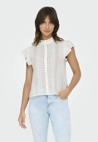 Blouse blanche en dentelle avec des manches volantes, des motifs verticaux et une fermeture à boutons. Assortie avec un jean en denim léger. Détails de tissu texturé visibles.