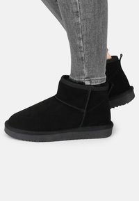 Botas de tobillo de ante negro con una puntera suave y redondeada y un perfil bajo. Presentan un puño plegable y una suela de goma texturizada para mayor tracción.