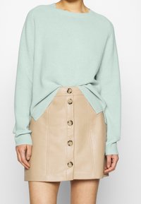 Pull en tricot vert menthe avec un col rond, associé à une jupe trapèze en cuir beige avec une fermeture à boutons à l'avant.