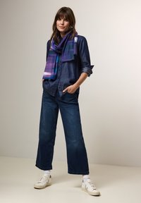 Denimskjorte med knaplukning og oprullede ærmer, parret med vide mørke jeans. Lilla tørklæde med en gradientdesign. Hvide sneakers.