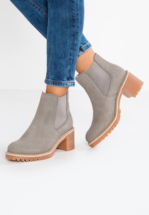 Grå ruskind ankelsstøvler med elastiske sidepaneler og brune kraftige hæle, båret med opsmøgede blå jeans på hvidt gulv.