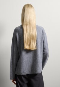 Grauer Wollpullover mit lockerer Passform, weiten Ärmeln und subtilen Kragenakzenten, kombiniert mit schwarzen Hosen. Weiche Textur und minimalistisches Design.