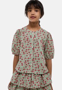 Blouse florale verte avec des manches bouffantes, ornée de fleurs rouges et roses et fermée par des boutons. L'ourlet froncé ajoute de la texture et du détail.