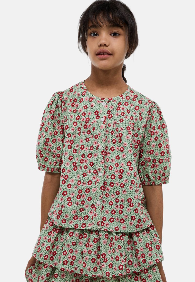 Blouse florale verte avec des manches bouffantes, ornée de fleurs rouges et roses et fermée par des boutons. L'ourlet froncé ajoute de la texture et du détail.