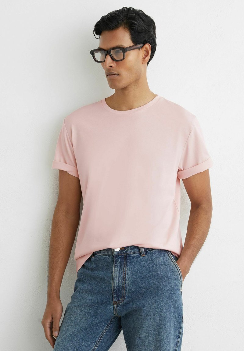 Férfi fekete szemüvegben, halvány rózsaszín, felhajtott ujjú pólóban és kék farmerral, egy kézzel a zsebében áll fehér fal előtt.