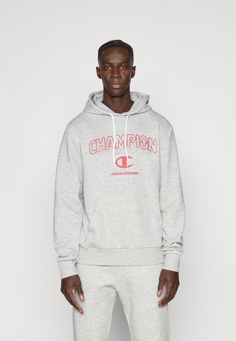Γκρι φούτερ με κουκούλα από μαλακό ύφασμα, με κόκκινο λογότυπο "CHAMPION" μπροστά και λευκά κορδόνια. Το μοντέλο φοράει ταιριαστό παντελόνι σε γκρι αποχρώσεις.