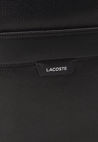 Juodas tekstūruotas odinis krepšys su išsiskiriančiu Lacoste logotipu. Su užtrauktuku ir lygiais akcentais kraštuose.