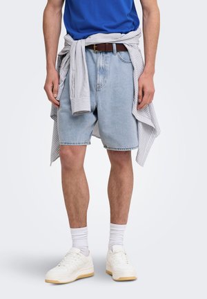 Homme portant un short en denim bleu clair, des chaussettes blanches, des baskets blanches, une chemise bleue et une chemise à manches longues rayée nouée autour de la taille.
