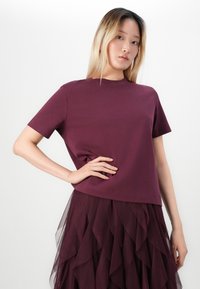 Young woman with blonde hair wears a burgundy short-sleeve top and layered tulle skirt, standing against a plain white background.