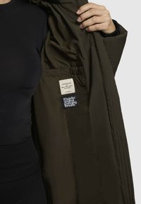 Olivově zelená zateplená bunda s quiltovanou texturou. Vnitřní štítky obsahují pokyny pro údržbu a název značky, viditelné na vnitřní podšívce.