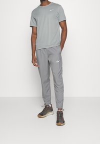 Camiseta atlética gris con cuello redondo, combinada con pantalones gris claro. Presenta el logo de Nike en blanco. Llevada con zapatillas deportivas oscuras.