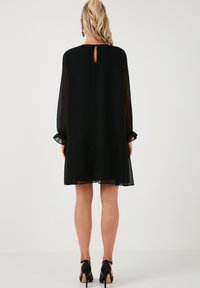 Black dress with sheer long sleeves, keyhole back detail, and ruffled cuffs, featuring a loose, A-line silhouette. Worn with heeled sandals.