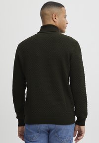 Mørkegrønn strikkegenser med et teksturert mønster, stående krage og ribbestrikkede mansjetter og kant. Kombinert med blå denimjeans.