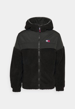 Zwarte gewatteerde jas met capuchon en ritssluiting, met een zachte fleece textuur en een glad bovenstuk op de schouders, voorzien van een klein rechthoekig logo in rood, wit en blauw op de borst.