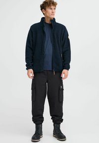 Dunkelblaues Fleece-Jackett mit Reißverschluss, strukturiertem Stoff. Kombiniert mit schwarzen Cargo-Hosen mit Taschen. Schwarze Schnürstiefel runden den Look ab.