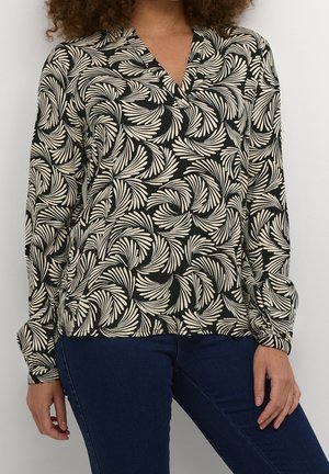 Woman wearing a black long-sleeve blouse with beige abstract leaf patterns and dark blue fitted jeans against a plain background.