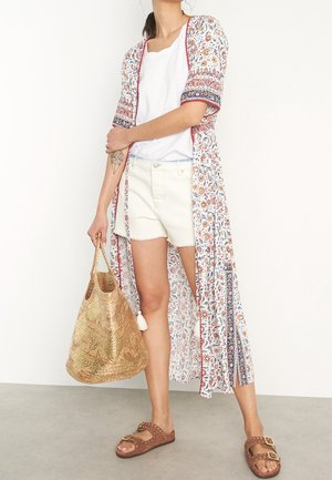 Kimono long floral en blanc, rouge et bleu, avec des manches courtes et des accents en franges, porté sur un haut blanc et un short en denim. Sandales marron.