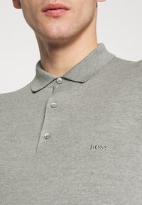 Polo gris côtelé avec un col à revers, doté de trois boutons et d'un logo "BOSS" brodé sur la poitrine. Texture de tissu lisse.
