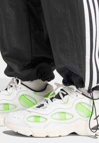 Vita sneakers med gröna detaljer och meshinformation. Rundad tå, tjock sula och snörning. Stilon med svarta träningsbyxor med vita ränder.