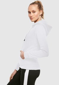 Weißer Hoodie mit einer Fronttasche, Kapuze mit Kordelzug und gerippten Bündchen. Schwarze Schriftzüge auf der Brust. Kombiniert mit schwarzen Hosen mit weißen seitlichen Streifen.