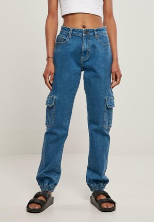 Pantalons cargo en denim bleu avec des poignets élastiques, poches avant et poches cargo sur le côté, associés à des sandales noires à bout ouvert.