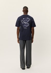 Μπλε ναυτικό βαμβακερό μπλουζάκι με λευκό γραφικό τύπωμα δύο φλυτζανιών καφέ και κείμενο στην πλάτη. Στρογγυλή λαιμόκοψη και κοντά μανίκια.