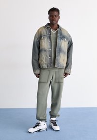 Dongerijakke med en falmet blå og beige gradient, lagd over en grønn topp. Olivengrønne joggebukser med store lommer og hvite sneakers.