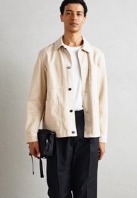Beige Baumwolljacke mit zwei Vordertaschen, Knopfverschluss und langen Ärmeln, getragen über einem weißen T-Shirt und kombiniert mit schwarzen Hosen.