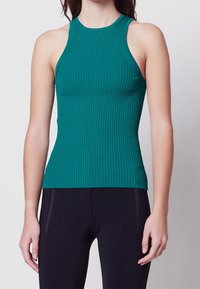 Mujer vistiendo una camiseta sin mangas de canalé en color teal y pantalones negros, de pie frente a un fondo claro y sencillo.