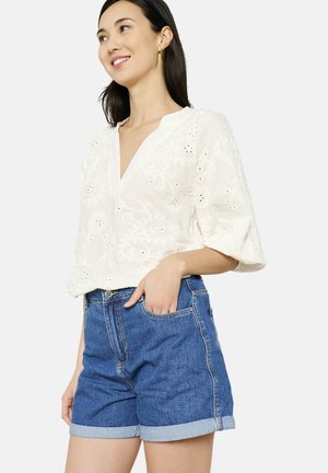 Blusa bianca ricamata con scollatura a V e maniche corte, abbinata a pantaloni shorts in denim a vita alta con orli arrotolati, di colore blu.