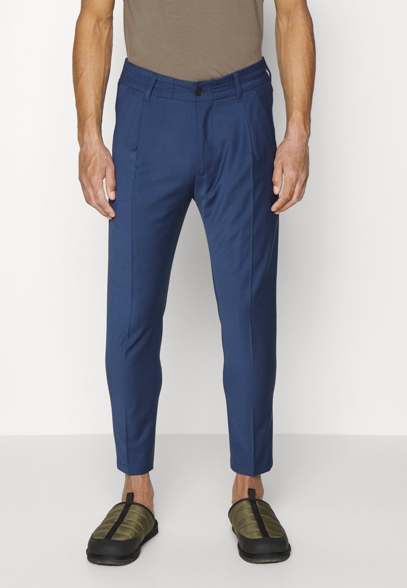 Navy blue tailored pants with a flat front design, side pockets, and a slim fit, paired with olive green slip-on shoes.