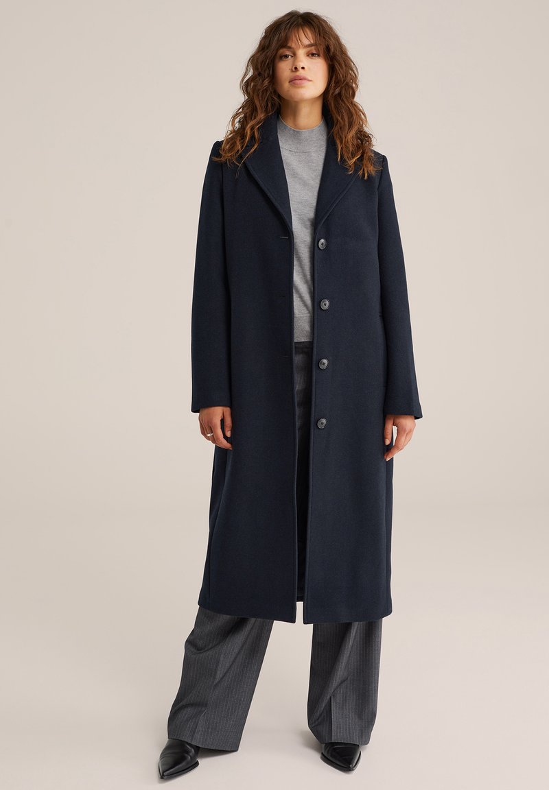 Kvinna med vågigt brunt hår, klädd i en lång mörkblå kappa, grå polo-tröja, grå randiga vida byxor och svarta spetsiga skor.