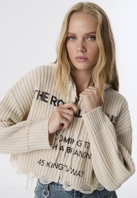 Haut à capuche beige coupé avec une texture côtelée, fermeture éclair à l'avant et détails en texte noir. L'ourlet à franges et les manches larges ajoutent des éléments de design uniques.