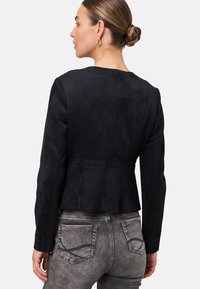 Donna con i capelli raccolti in un bun, che indossa orecchini a cerchio dorati, una giacca nera aderente a maniche lunghe e jeans in denim grigio sbiadito, di spalle.