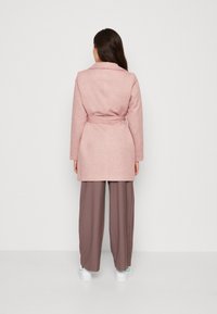 Una donna con lunghi capelli scuri indossa un cappotto rosa con cintura lungo fino al ginocchio, pantaloni larghi marroni e sneakers bianche, in piedi di spalle su uno sfondo neutro.