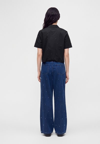 Femme aux longs cheveux foncés portant un t-shirt noir à manches courtes et un jean bleu à jambes larges avec des motifs subtils de petits points blancs, debout, de dos.