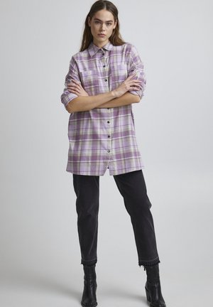 Herenmode-geïnspireerde geruite shirt in paarse en groene tinten, met knoopsluiting aan de voorkant, lange mouwen opgerold, gecombineerd met zwarte denim en haklaarzen.