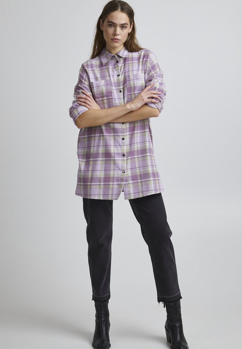 Herenmode-geïnspireerde geruite shirt in paarse en groene tinten, met knoopsluiting aan de voorkant, lange mouwen opgerold, gecombineerd met zwarte denim en haklaarzen.