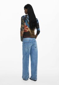 Langärmliges Hemd mit einem bunten floralen und abstrakten Design; kombiniert mit weit geschnittenen blauen Jeans. Strukturiertem Stoff mit unterschiedlichen Farbtönen.