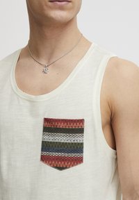 Cămașă fără mâneci de culoare bej des, având un buzunar cu model în dungi roșii, verzi, gri și albastre. Guler rotunjit, textură moale.